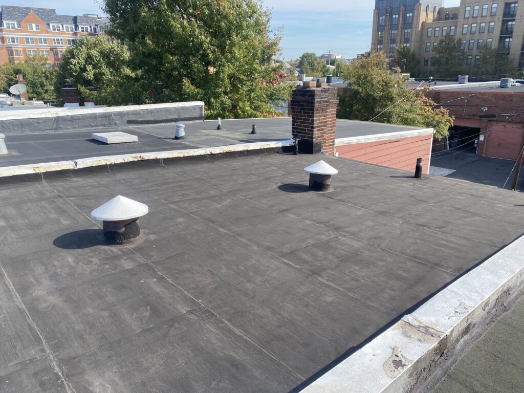 Worn low-slope flat roof with vent stacks and a brick chimney before a torch-down replacement in Alexandria, Virginia