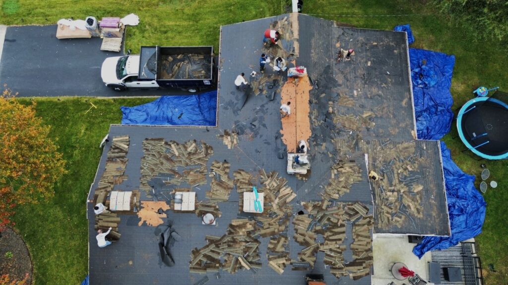 Aerial view of the roof mid-tear-off, with a dump truck, staged shingle bundles and landscaping tarps visible