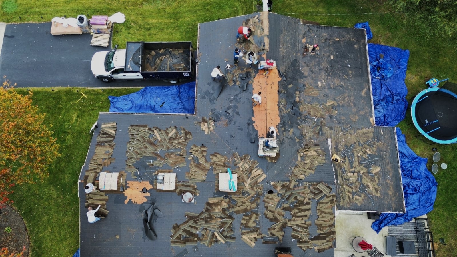 Aerial view of the same roof mid-tear-off, with a dump truck, staged shingle bundles and landscaping tarps visible
