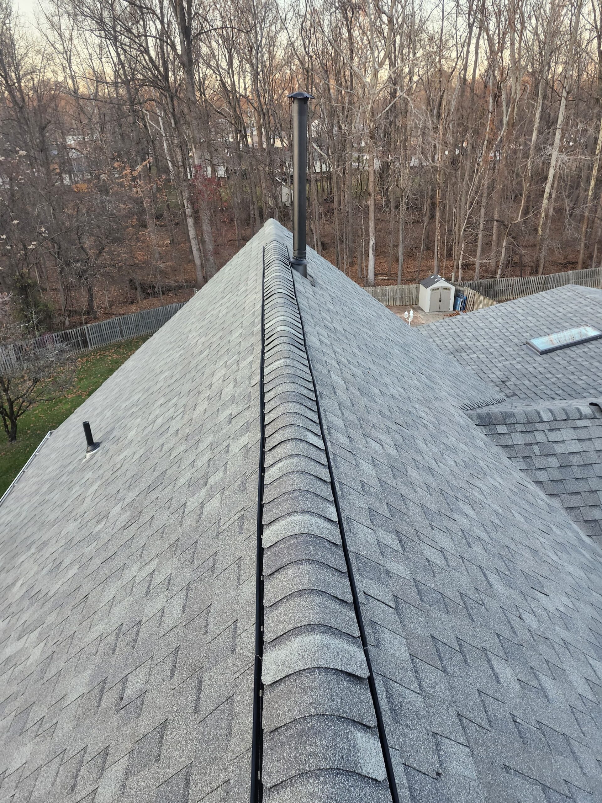 Finished dark gray architectural shingle roof ridge, with the same wooded backdrop