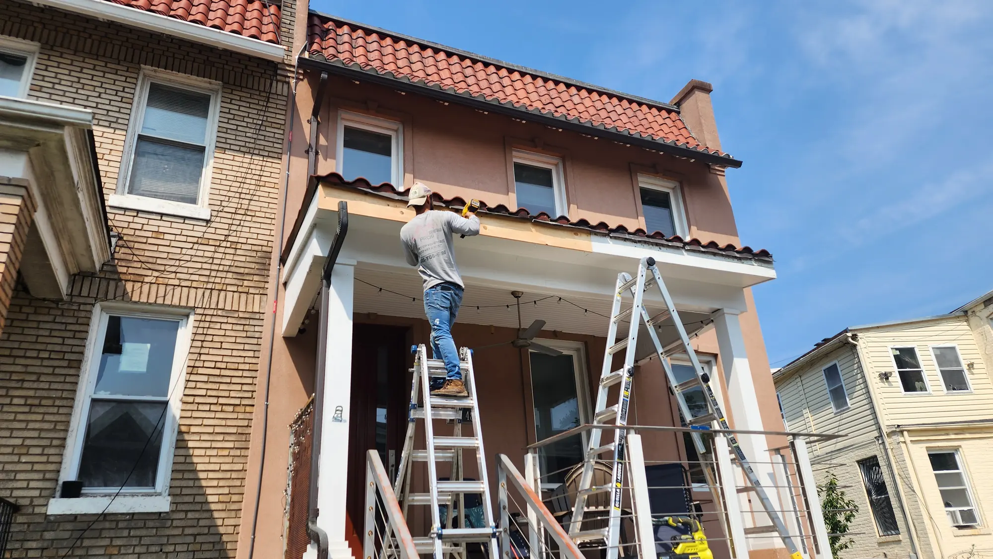 Roof maintenance on a Washington, D.C. row house, with a crew member on a ladder working at the gutter and fascia line