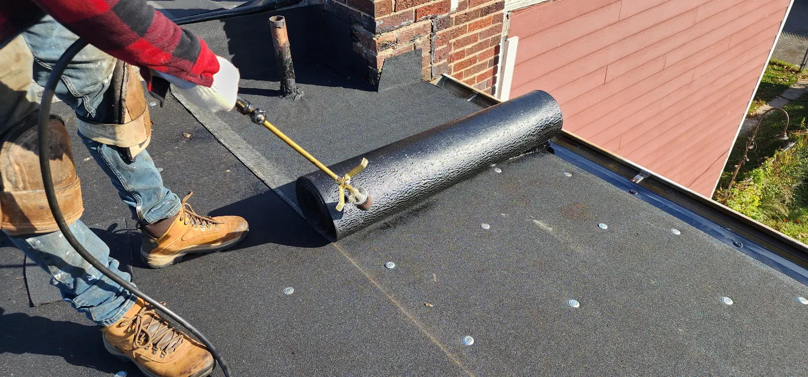 Roofer unrolling and torching a roll of modified-bitumen membrane across a prepared roof deck